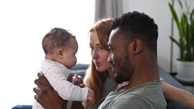 Young multiracial parents enjoy time together with newborn at cozy home. An African dad and caucasian mom gently holding sweet baby girl and caress her with love