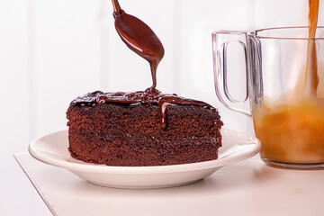 Piece of cake on white plate. pouring chocolate from spoon, cuo of coffee with milk