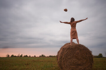 Sunset in a field. Heavy rainy clouds  flow away leaving space for sun. Woman stays on a haystack and tosses a hat up. She is in safari dress.