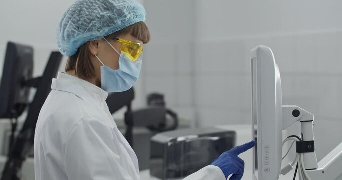Female young researcher in protective mask and yellow googles working on computer. Woman scientist in white rob and bouffant cap working with special equipment at modern clinic.