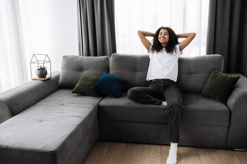 A beautiful young woman is relaxing on the couch in living room at home, smiling. African american girl takes a break from online distant work, or have lazy leisure. Happy attractive female dreaming