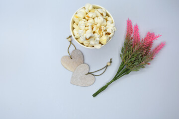 Fototapeta na wymiar From above selective focus of small light beige wooden hearts next to popcorn in bright disposable cup and bouquet of pink delicate flowers with tiny green leaves in light of lamps on white background