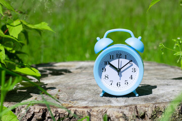 Sunlit blue alarm clock on wooden cut stump with green bokeh background. Metaphor or concept of summer season coming.