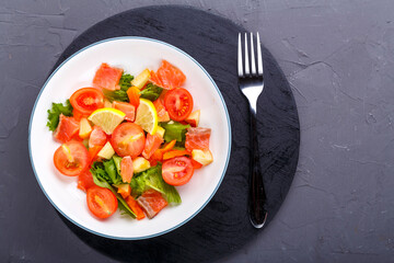 Salad with cold salmon and cherry tomatoes in a plate on a concrete background with a black stand next to a fork.