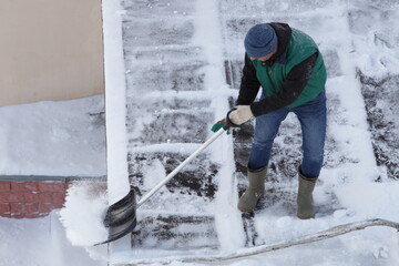 A worker man without a safety belt and helmet cleans snow from the roof with a shovel on a winter day