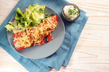 Pointed bell pepper stuffed with rice, tuna, tomatoes and cheese on a blue plate with lettuce salad, and dip, on a napkin and a bright wooden table, copy space, high angle view from above