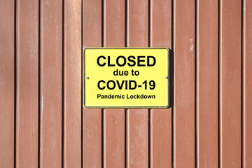 Shuttered wooden gate with a yellow sign and text Closed due to Covid-19 Pandemic Lockdown, many retailers are forced to do this during the coronavirus crisis, copy space
