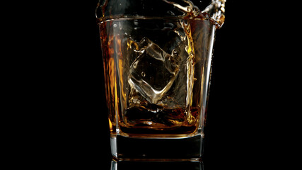 Glass of whisky with splashing liquid and ice rocks inside.