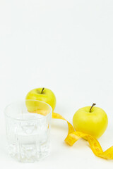 A glass of water and a measuring tape wrapped around two green apples as a symbol of diet. The concept of a healthy lifestyle, food and sports, on a white background. Side view with place for text or 