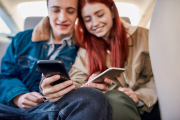 Close up of hands of two teenagers smiling, using their smartphones while sitting together on back seat in the car