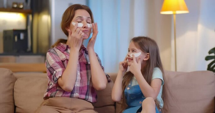 Happy mother and daughter sitting together on couch doing under eyes mask