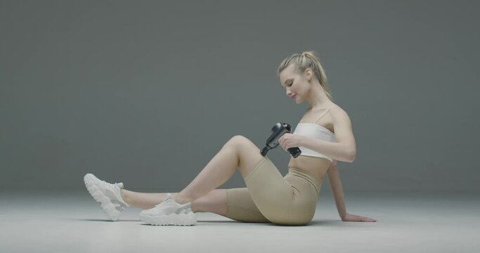 Athletic young blond girl massaging leg by handheld percussion massager on gray studio backdrop for sore muscle relief