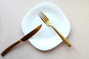 White plate with a golden fork and knife on a gray table, top view, close-up