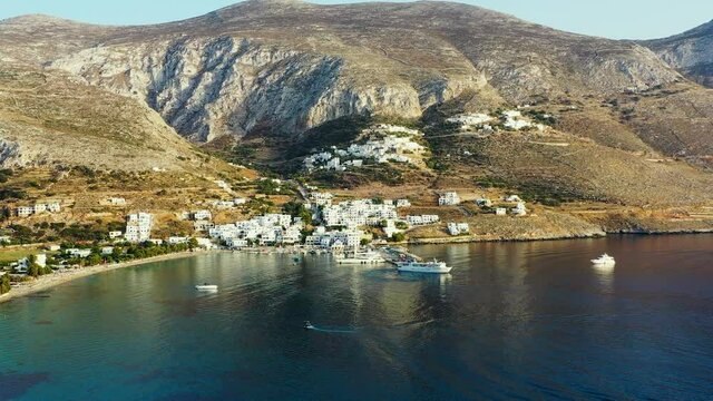 Drone video of Aegialis in Greece. Drone fly high over the small town, surrounded by mountains, and clear blue water. There is a small marina, and there is a boat sailing towards the harbour. 1080p