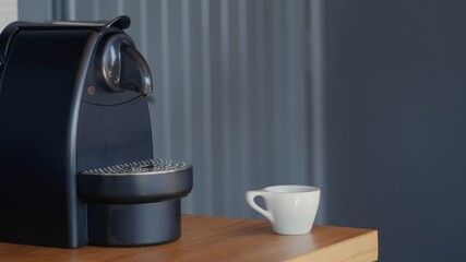 Black capsule espresso coffee machine and small white ceramic cup on a light wood table. Grey background. High-quality jpg photo