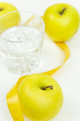 A glass of water and a measuring tape wrapped around three green apples as a symbol of diet. The concept of a healthy lifestyle, food and sports, on a white background. side view