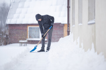 Young adult man hands holding blue shovel and shoveling fresh white snow at yard of house during...