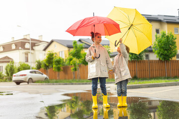 Two happy sisters in yellow rubber rain boots play with bright umbrellas in the street after the rain