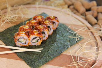 Japanese sushi roll with unagi eel, cucumber wrapped in rice with orange flying fish roe Tobiko (Masago) served with sauce on nori seaweed sheet. Wooden board on background, chopsticks on foreground
