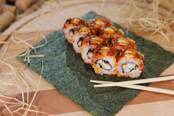 Japanese sushi roll with unagi eel, cucumber wrapped in rice with orange flying fish roe Tobiko (Masago) served with sauce on nori seaweed sheet. Wooden board on background with chopsticks near dish
