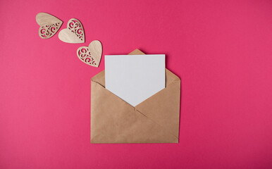 Craft envelope with a blank sheet of paper inside and wooden hearts on the hot pink background. Romantic love letter for the Valentine's day concept. Space for text.