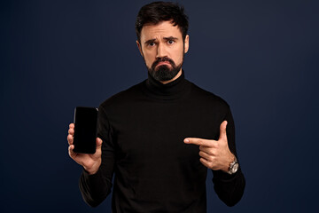 Technology, people and communication concept.Portrait of upset and disappointed,gloomy bearded caucasian man pointing finger at mobile phone display and grimacing unsatisfied, Pacific Blue background