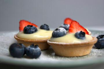Macro shot of French dessert. Little tarts with custard, fresh berries and powdered sugar. Tartlets with strawberries, blueberries served with icing sugar (castor sugar) on gray and white plates
