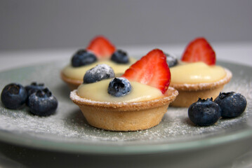 Side view of three French desserts. Little tarts with custard, fresh berries and powdered sugar. Tartlets with strawberries, blueberries served with icing sugar (castor sugar) on gray and white plates