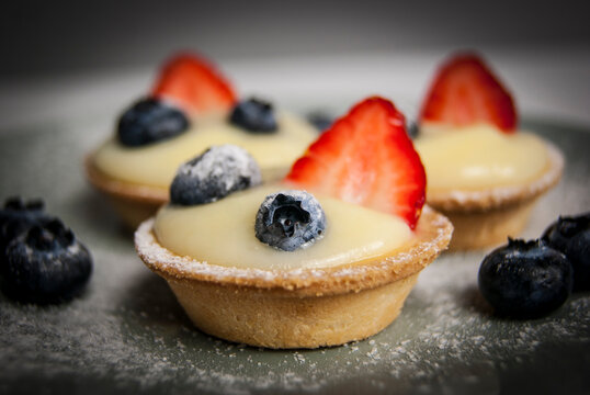 Side view of three French desserts. Little tarts with custard, fresh berries and powdered sugar. Tartlets with strawberries, blueberries served with icing sugar (castor sugar) on gray and white plates