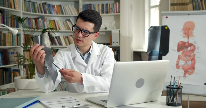 Male doctor holding x ray photo of chest while having online medical consultation in office.Young man in glasses and white rob using laptop while communicating with patient