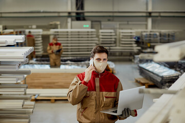 Male worker using laptop while talking on the phone at carpentry workshop.