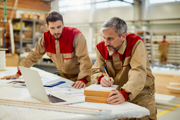Male worker writing notes while using laptop with colleagues at carpentry workshop.