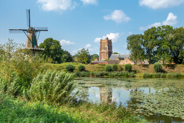 The historical town of Woudrichem, The Netherlands, with a windmill and church, seen from behind a pond with green shrubs on a sunny day