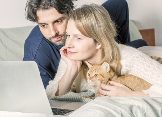 Young couple relaxing on bed with their cat using laptop to video call  their family. Soft focus laptop. Love, happiness, technology, people and fun