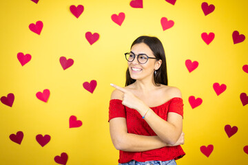 Young beautiful woman over yellow background with red hearts confused and pointing with hand and finger to the side