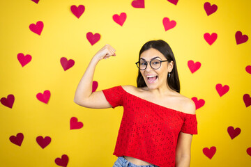 Obraz na płótnie Canvas Young beautiful woman over yellow background with red hearts showing arms muscles smiling proud