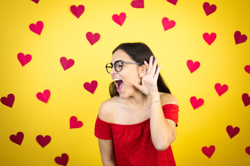 Young beautiful woman over yellow background with red hearts surprised with hand over ear listening an hearing to rumor or gossip