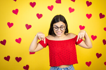 Young beautiful woman over yellow background with red hearts surprised, looking down and pointing down with fingers and raised arms
