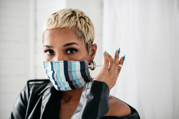 Beautiful young Black woman with short blond hair wearing mask manicure blue nails holding and applying mask to face