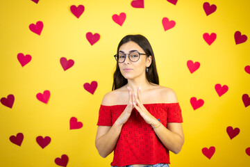 Young beautiful woman over yellow background with red hearts with Hands together and fingers crossed smiling relaxed and cheerful. Success and optimistic