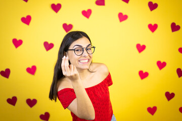 Young beautiful woman over yellow background with red hearts making capice or money gesture, telling you to pay your debts!