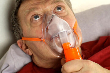 Sick senior adult man breathing through oxygen mask, rolling his eyes. Treatment of asthma, allergies, bronchitis, pneumonia in Covid-19 and respiratory diseases. Selective focus. Close-up. Indoor. - 405630709