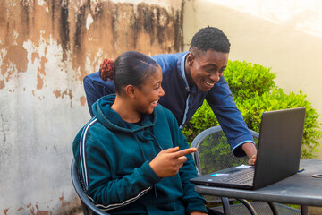young black man and lady happy together operating a laptop while sitting outdoor. - obrazy, fototapety, plakaty