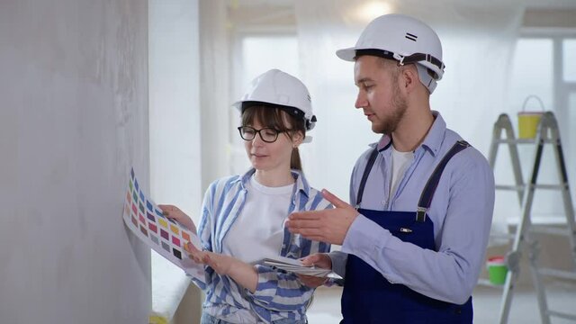 male painter and female designer in hard hats discuss wall paint from a color palette during interior finishing work on a construction site