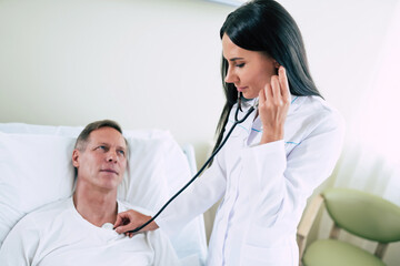 Young modern confident woman doctor in a white coat with a stethoscope is examining the patient's feelings while he is lying on the bed in the hospital ward.