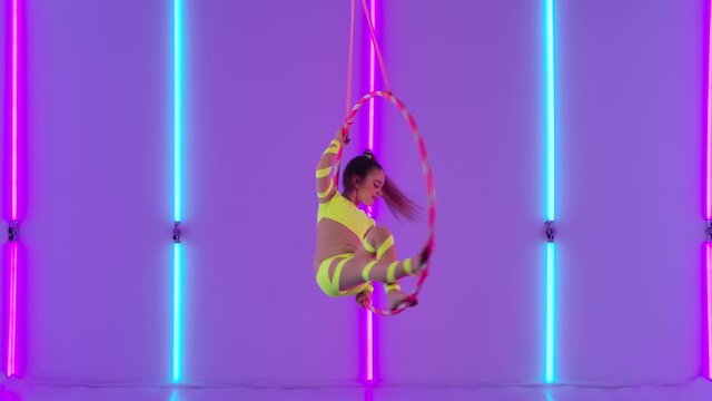 Young girl aerial acrobat rotates in the air on a hoop. Circus performer in a yellow leotard against the background of bright neon lights in the studio. Slow motion.