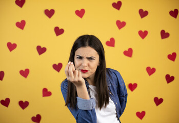 Young caucasian woman over yellow background with red hearts making capice or money gesture, telling you to pay your debts!