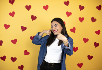 Young caucasian woman over yellow background with red hearts doing the “call me” gesture with her hands.