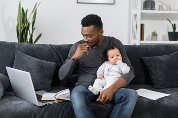 A thoughtful freelancer, student dad with an infant sit on the couch, he works from home. A young African-American father using laptop for remote work, while looking after his cute baby daughter