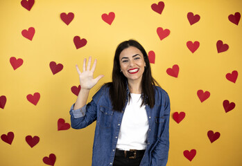 Young caucasian woman over yellow background with red hearts showing and pointing up with fingers number five while smiling confident and happy